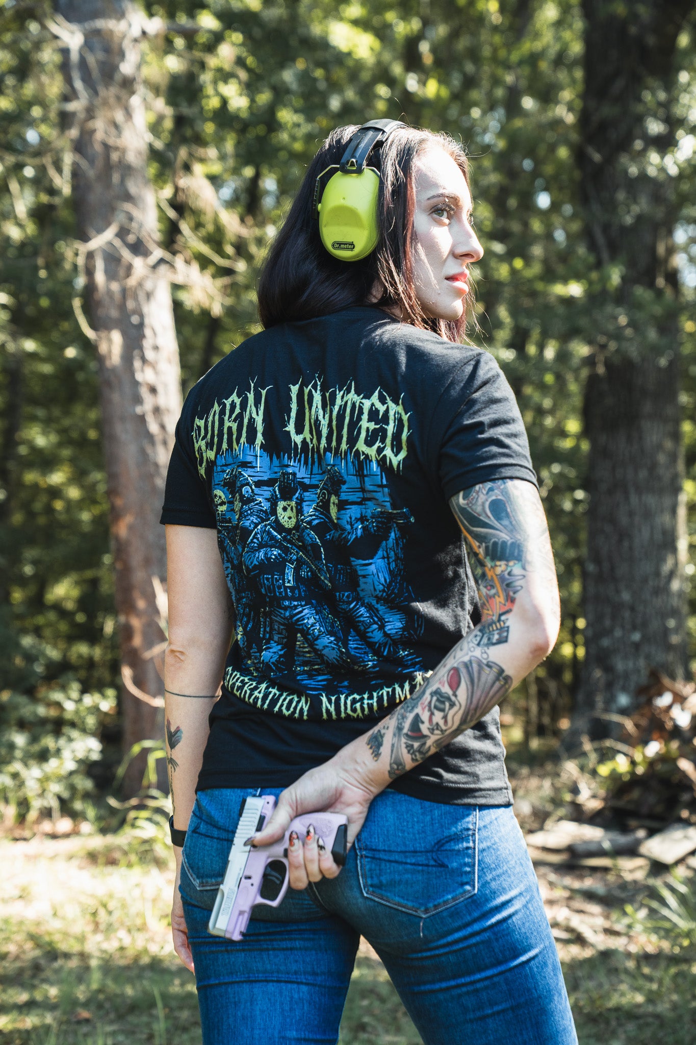Seen from behind, a person wears a black Born United Private Label Operation Nightmare Tee - Glow in the Dark with tactical figures and neon lettering while holding a pink pistol at a wooded firing range.