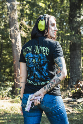 Seen from behind, a person wears a black Born United Private Label Operation Nightmare Tee - Glow in the Dark with tactical figures and neon lettering while holding a pink pistol at a wooded firing range.