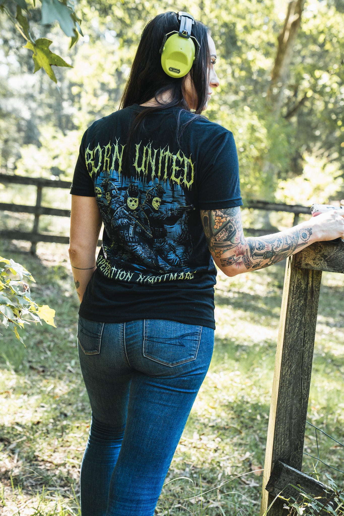 A woman wears a black Born United Private Label Operation Nightmare Tee - Glow in the Dark, featuring a graphic of soldiers in hockey masks on the back, standing by a wooden fence.