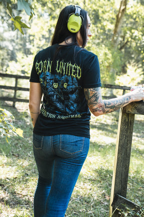 A woman wears a black Born United Private Label Operation Nightmare Tee - Glow in the Dark, featuring a graphic of soldiers in hockey masks on the back, standing by a wooden fence.