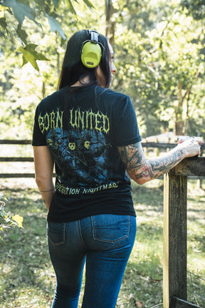 A woman wearing a black Operation Nightmare Tee - Glow in the Dark, featuring masked figures holding firearms, stands at a wooden fence while wearing lime green ear protection.