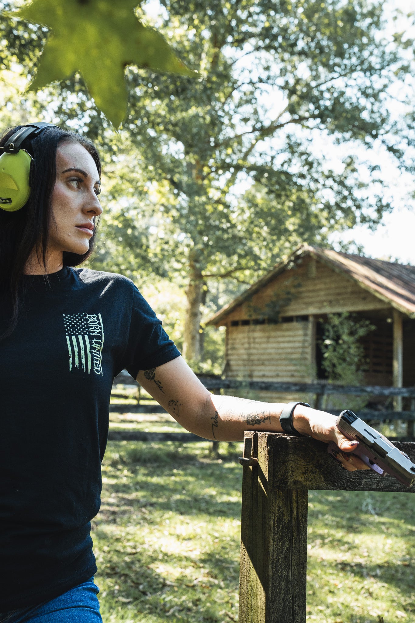A woman in premium cotton hearing protection and a black Operation Nightmare Tee - Glow in the Dark stands in a wooded area, resting her hand on a wooden post while holding a handgun.