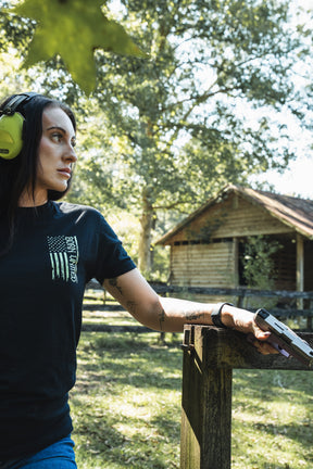 A woman in premium cotton hearing protection and a black Operation Nightmare Tee - Glow in the Dark stands in a wooded area, resting her hand on a wooden post while holding a handgun.