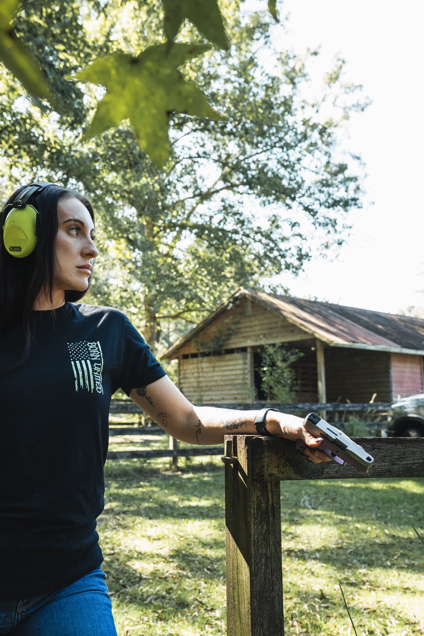 A woman wearing hearing protection and a black Operation Nightmare Tee - Glow in the Dark stands by a wooden fence, holding a handgun.