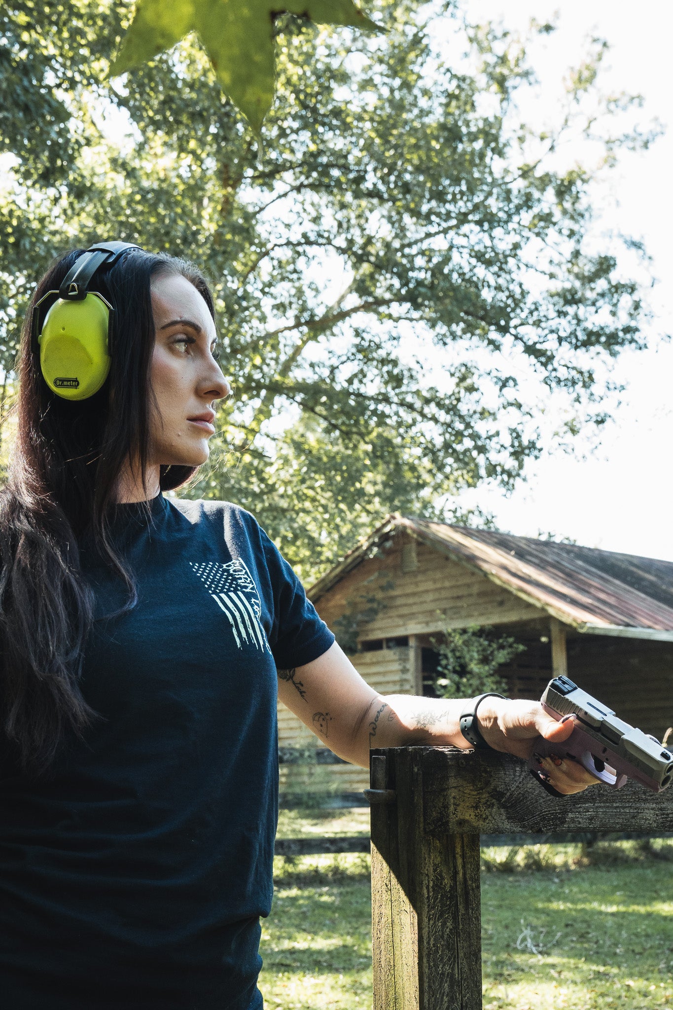 A woman wearing ear protection and an Operation Nightmare Tee - Glow in the Dark cotton t-shirt with an American flag graphic stands outdoors holding a handgun resting on a wooden fence.