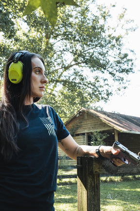A woman wearing ear protection and an Operation Nightmare Tee - Glow in the Dark cotton t-shirt with an American flag graphic stands outdoors holding a handgun resting on a wooden fence.