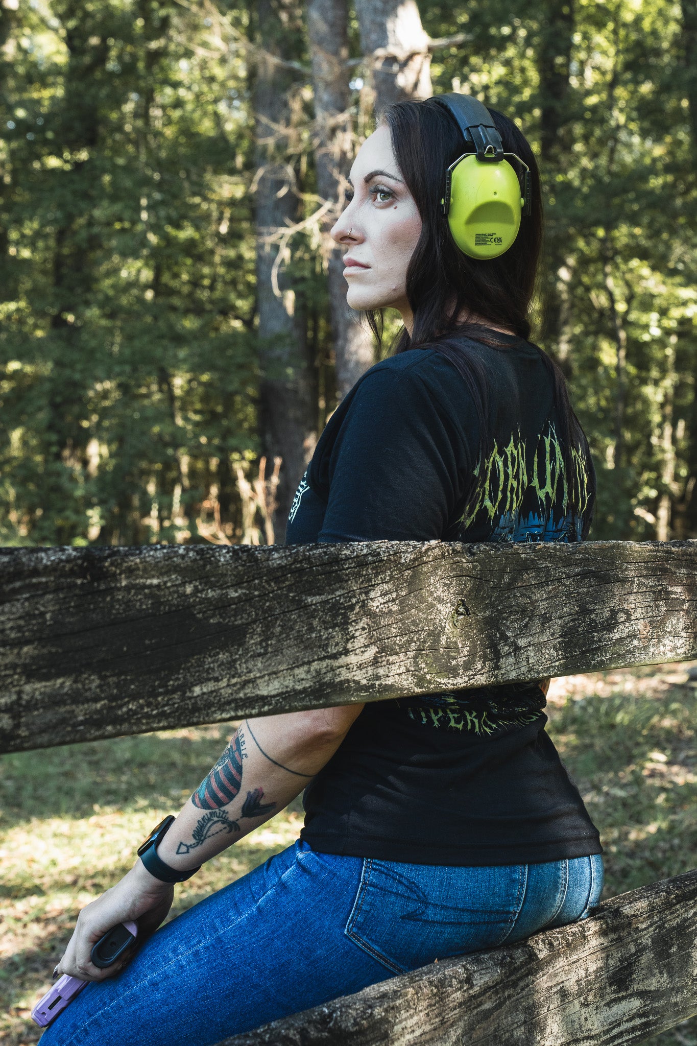 A woman wearing a black Operation Nightmare Tee - Glow in the Dark cotton t-shirt and green ear protection sits on a wooden fence outdoors.
