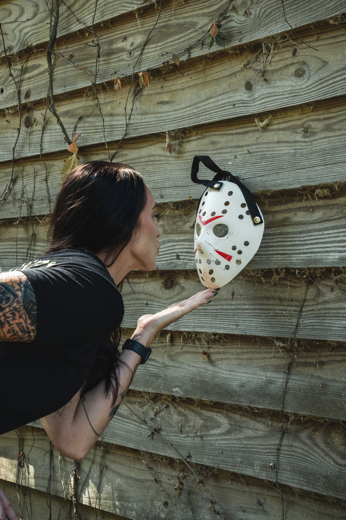A woman wearing a black Operation Nightmare Tee - Glow in the Dark blowing a kiss toward a hockey-style mask mounted on a wooden wall.