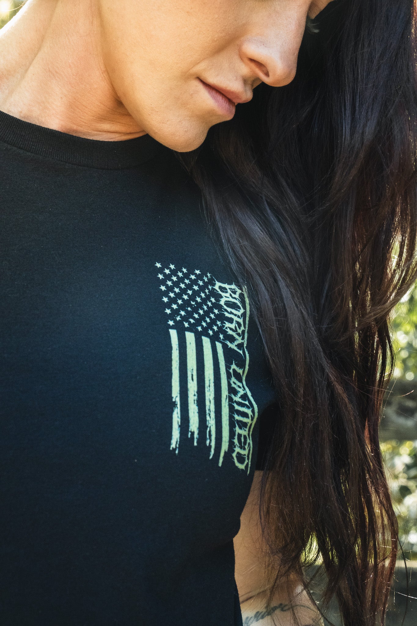 A person wears a black Operation Nightmare Tee - Glow in the Dark, featuring a vertical American flag graphic with the text BORN UNITED integrated into the stripes.