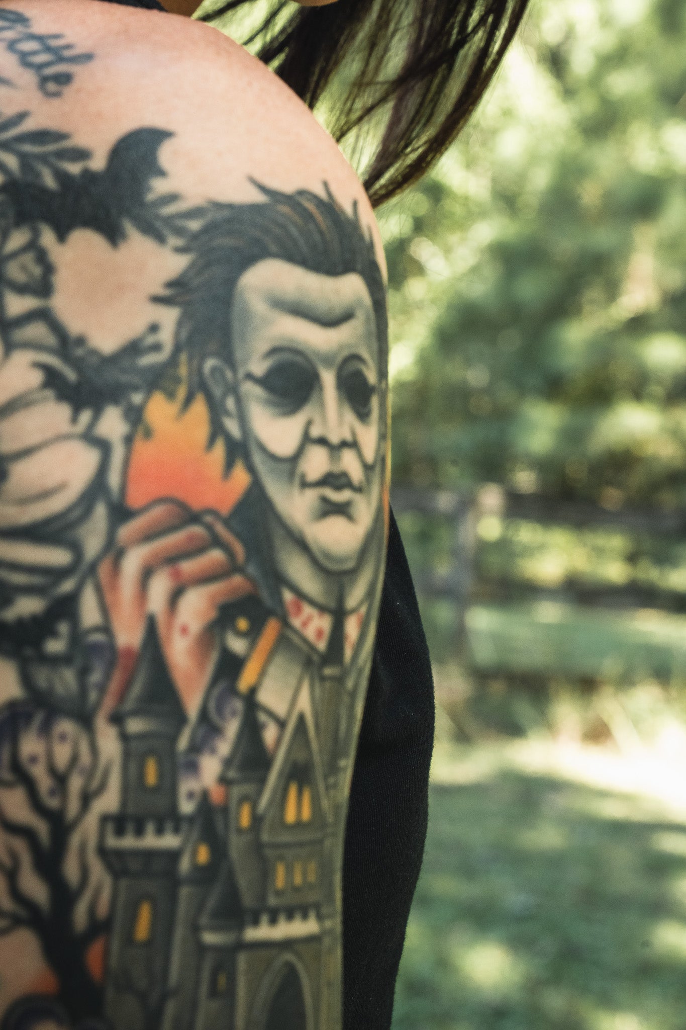 A person’s upper arm wearing the Operation Nightmare Tee - Glow in the Dark, featuring a glow-in-the-dark horror character tattoo above a dark castle and forest on premium cotton.