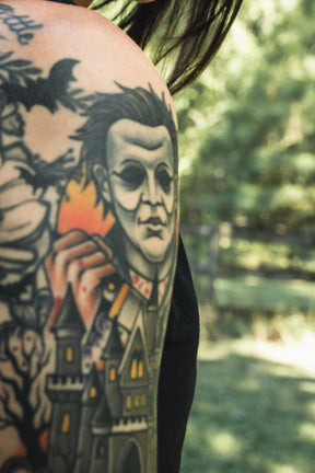 A person’s upper arm wearing the Operation Nightmare Tee - Glow in the Dark, featuring a glow-in-the-dark horror character tattoo above a dark castle and forest on premium cotton.