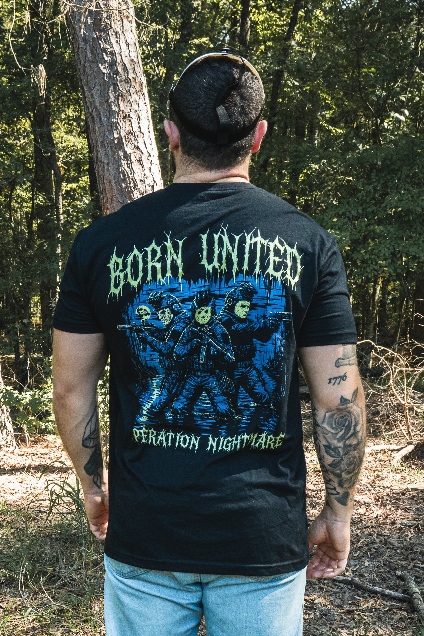 Back view of a man wearing a black Operation Nightmare Tee - Glow in the Dark featuring three masked tactical figures with BORN UNITED and OPERATION NIGHTMARE text.