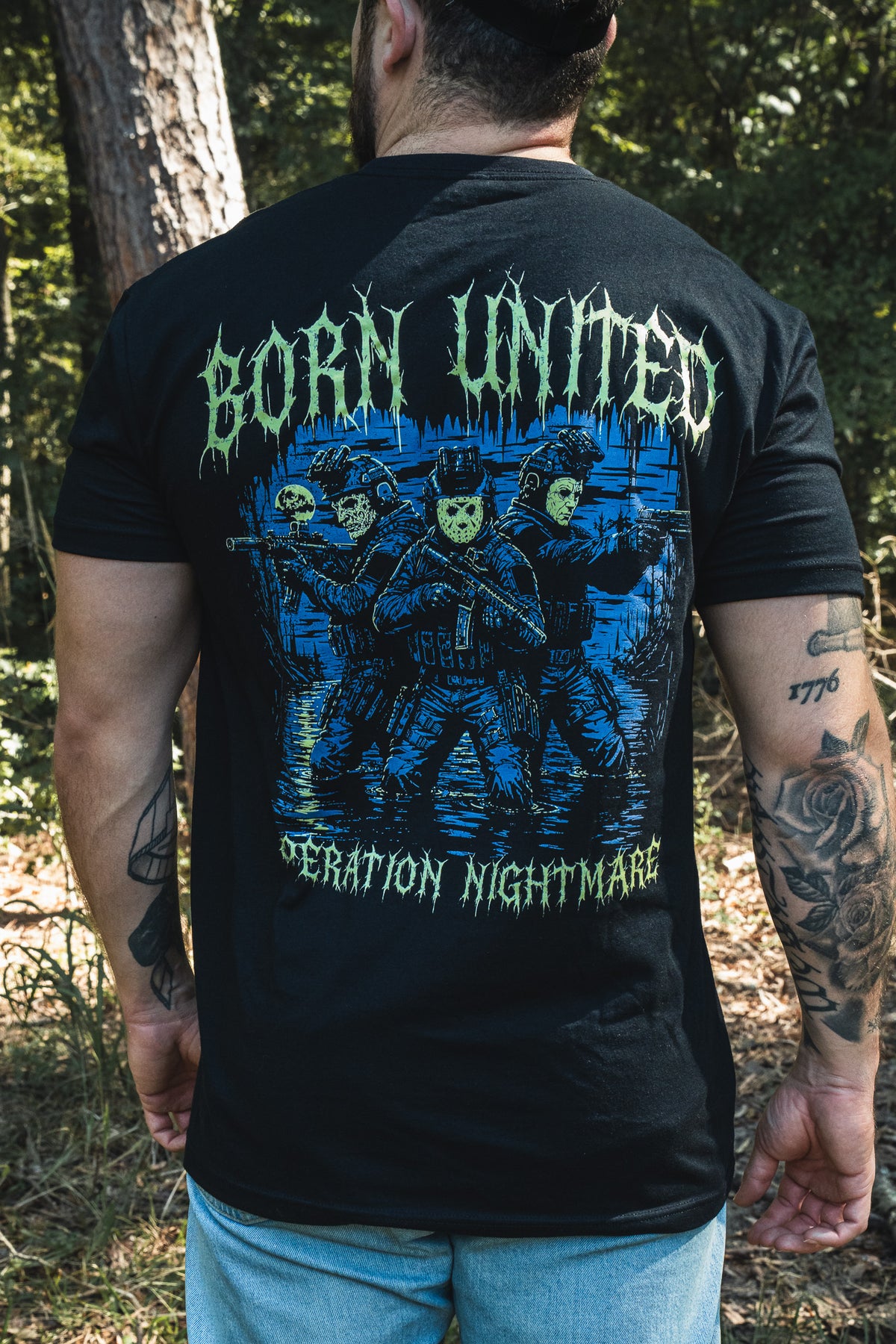 A person wearing a premium cotton black Operation Nightmare Tee - Glow in the Dark, featuring a graphic of three figures in tactical gear and hockey masks in water with the text BORN UNITED.