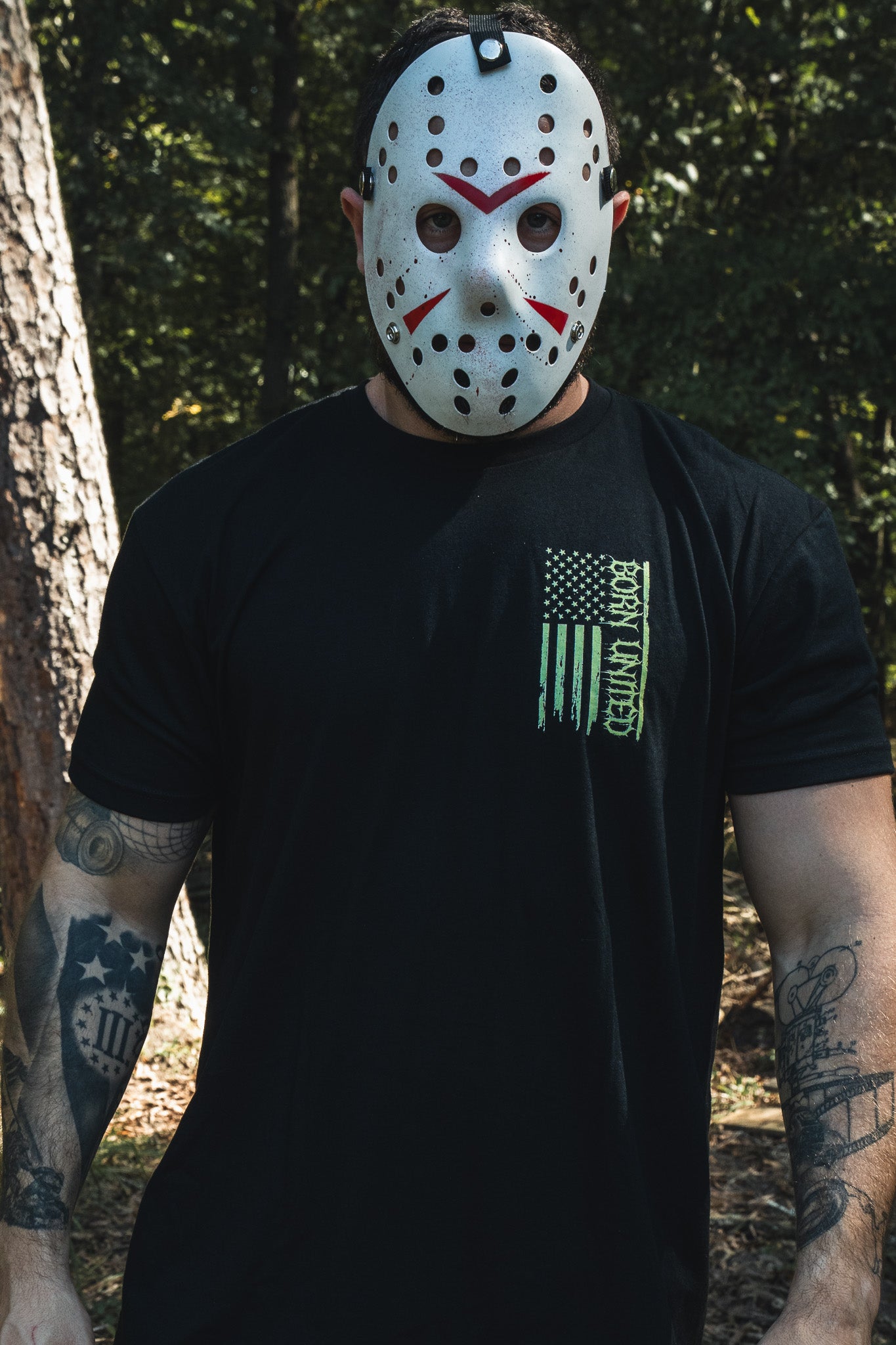 A man in a hockey mask wears a black Operation Nightmare Tee - Glow in the Dark, featuring a green, vertical American flag graphic.