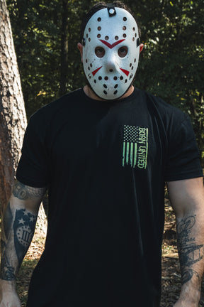 A man in a hockey mask wears a black Operation Nightmare Tee - Glow in the Dark, featuring a green, vertical American flag graphic.