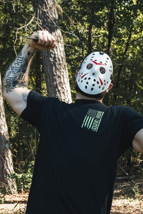 A person wearing a white hockey mask and the Born United Private Label Operation Nightmare Tee - Glow in the Dark, posing in a forest while holding a small knife.