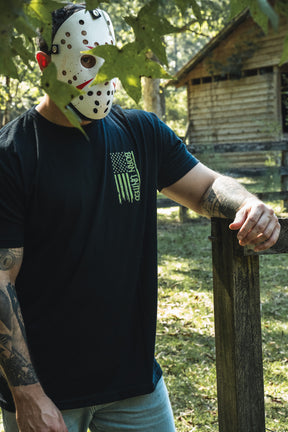A person stands in a wooded area wearing a Born United Private Label Operation Nightmare Tee - Glow in the Dark, a black cotton t-shirt featuring a green graphic of a distressed American flag and hockey mask.