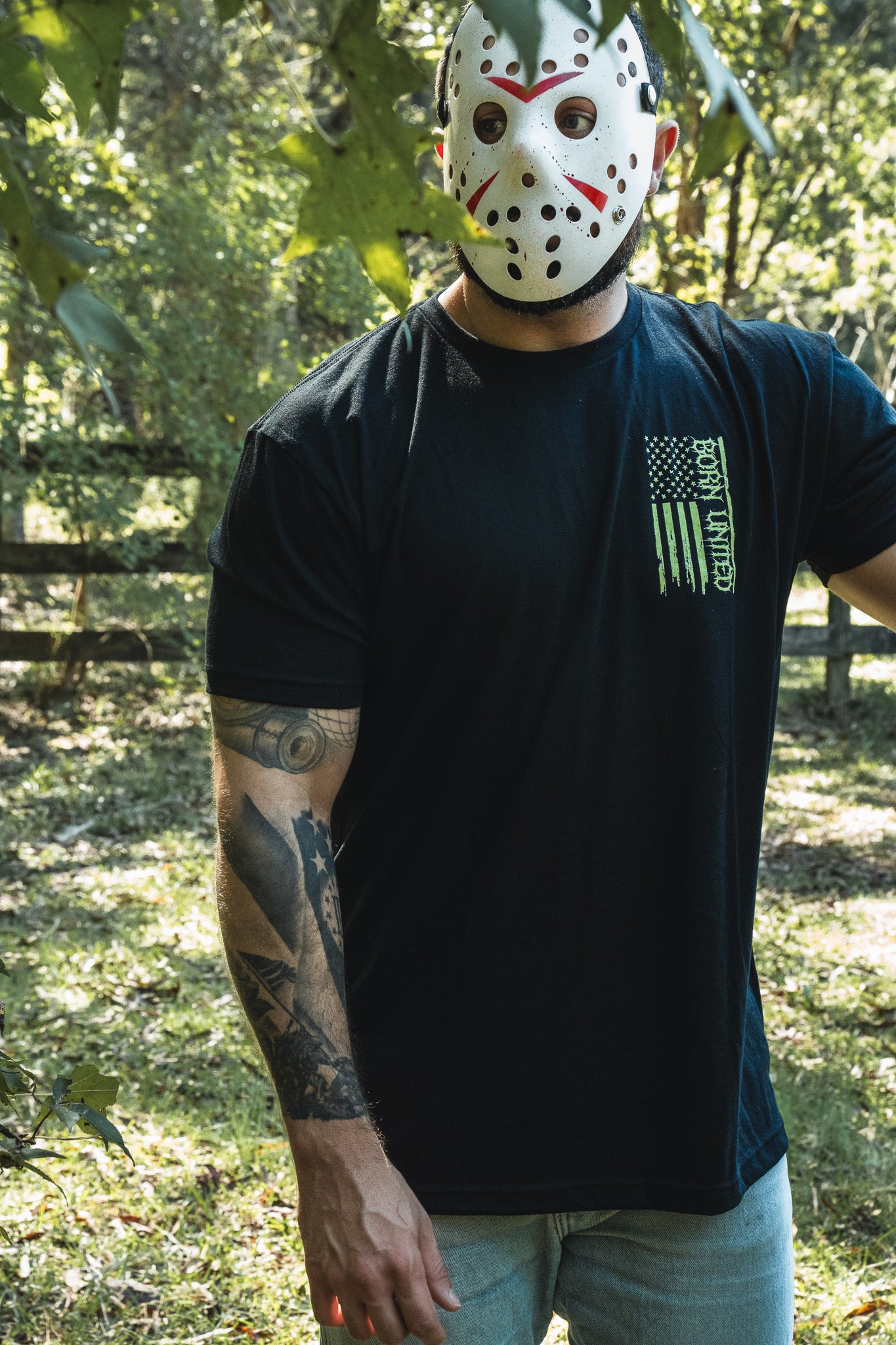 A person wearing a hockey mask and an Operation Nightmare Tee - Glow in the Dark, a black premium cotton T-shirt featuring a green, vertically oriented American flag graphic.