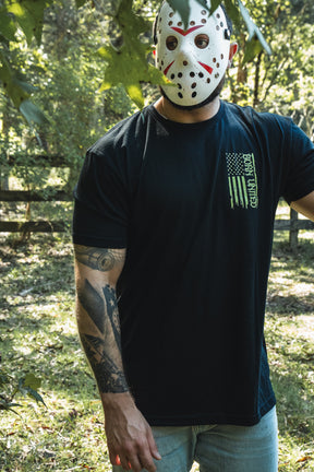 A person wearing a hockey mask and an Operation Nightmare Tee - Glow in the Dark, a black premium cotton T-shirt featuring a green, vertically oriented American flag graphic.