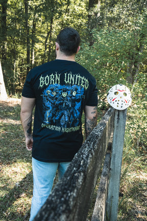 A man wears an Operation Nightmare Tee - Glow in the Dark featuring soldiers in hockey masks, standing near a matching mask resting on a wooden forest fence post.
