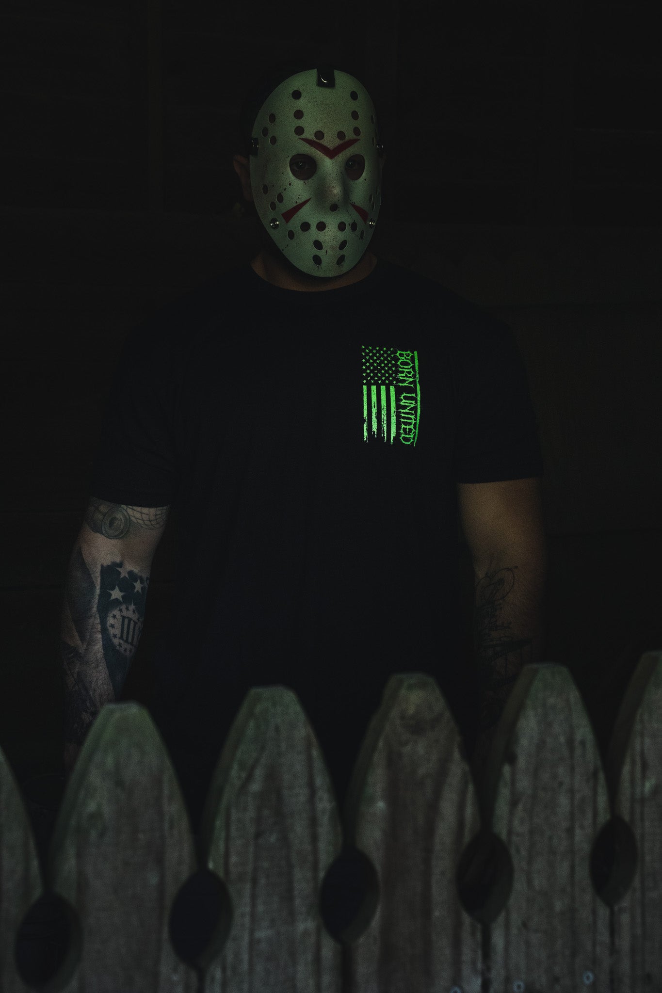 A person wearing a hockey mask and an Operation Nightmare Tee - Glow in the Dark, featuring a glowing green vertical American flag graphic.