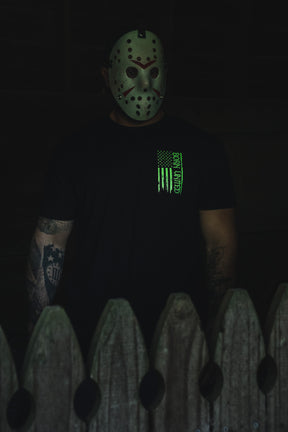 A person wearing a hockey mask and an Operation Nightmare Tee - Glow in the Dark, featuring a glowing green vertical American flag graphic.