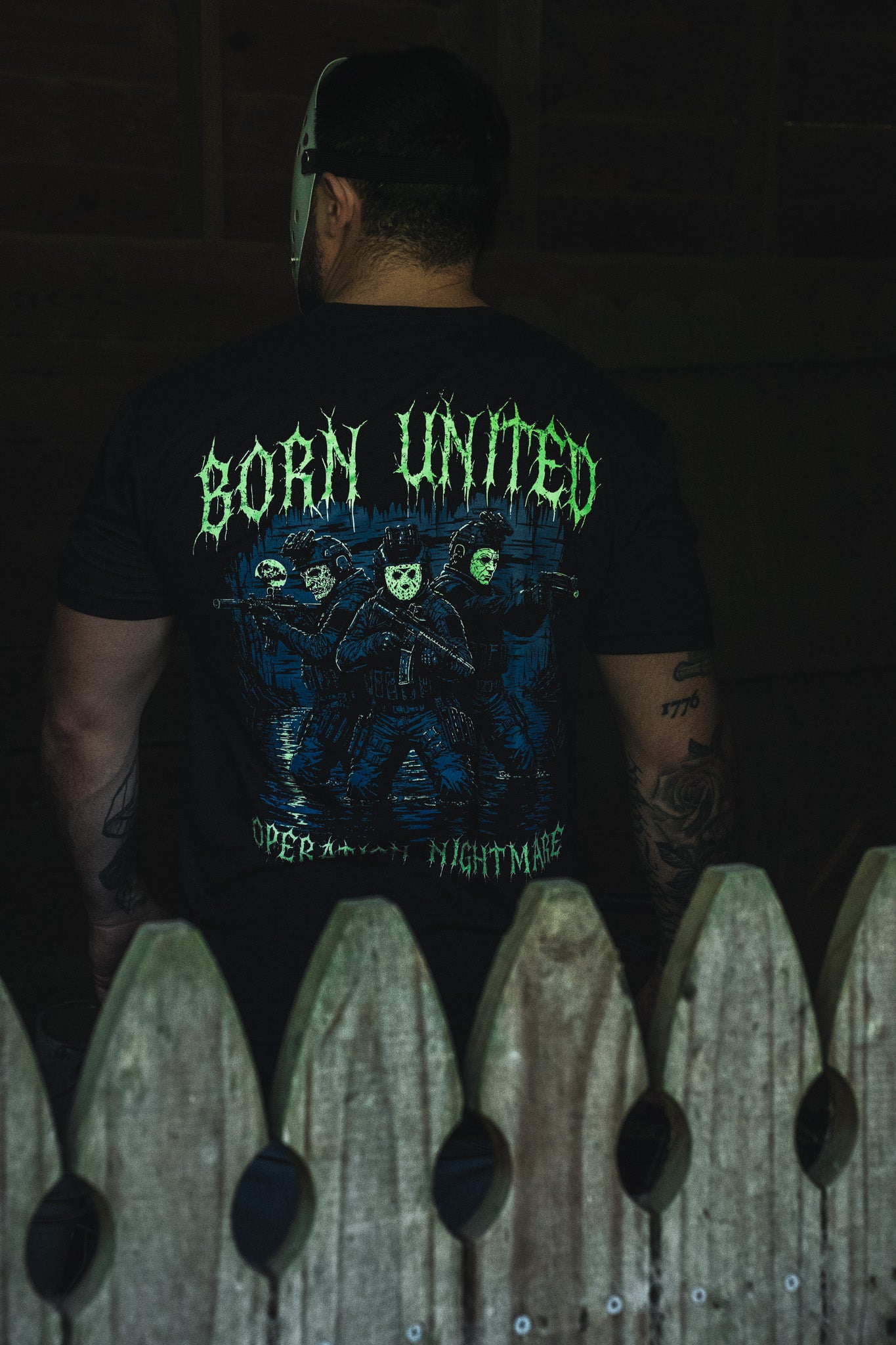 A person wears a black Operation Nightmare Tee - Glow in the Dark, featuring a glow-in-the-dark graphic of three figures in tactical gear, including one in a hockey mask, with the text Born United.