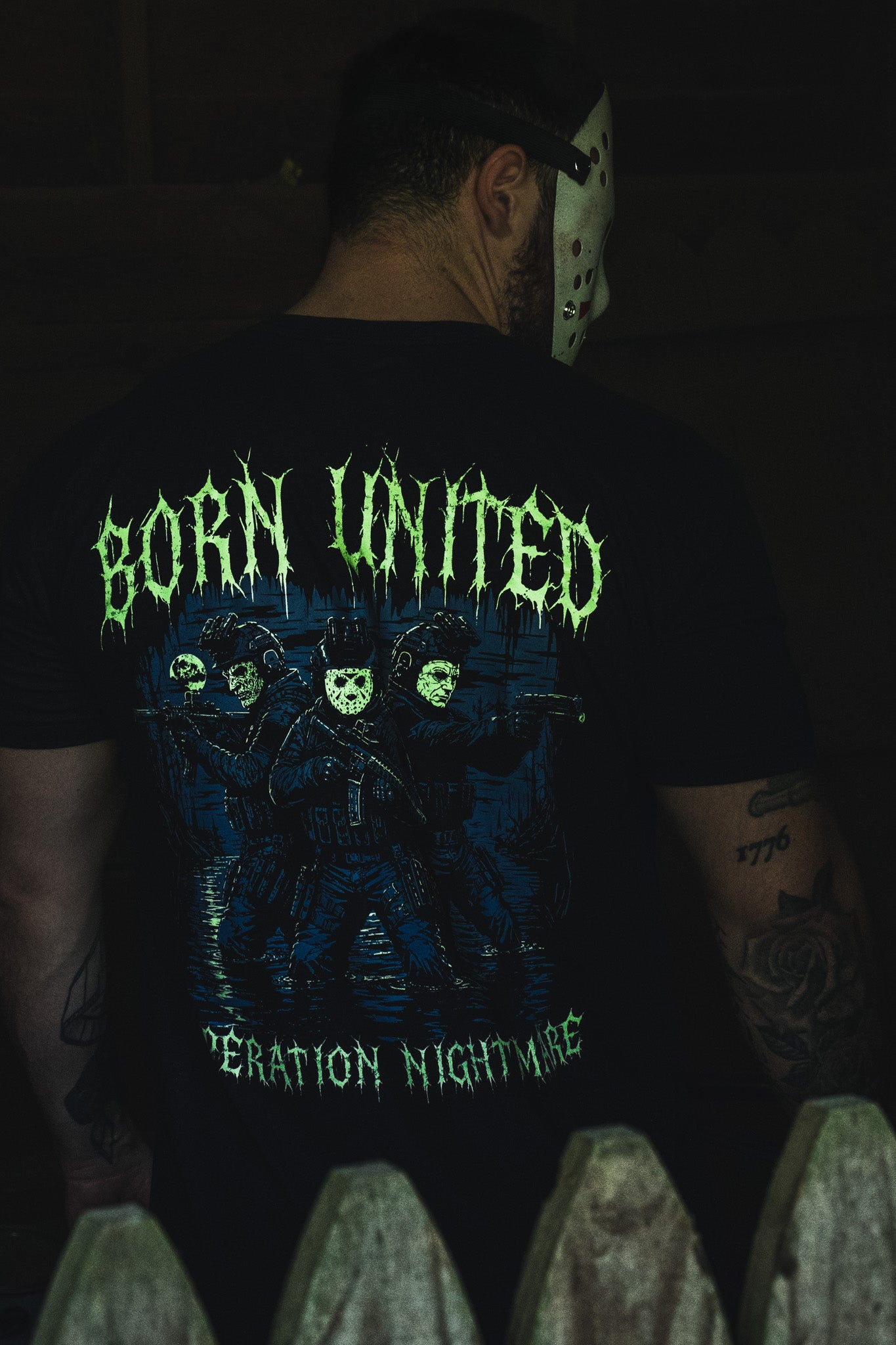 A person wears a black Operation Nightmare Tee - Glow in the Dark, featuring a glow-in-the-dark graphic of three armed figures in hockey masks with drippy green lettering.