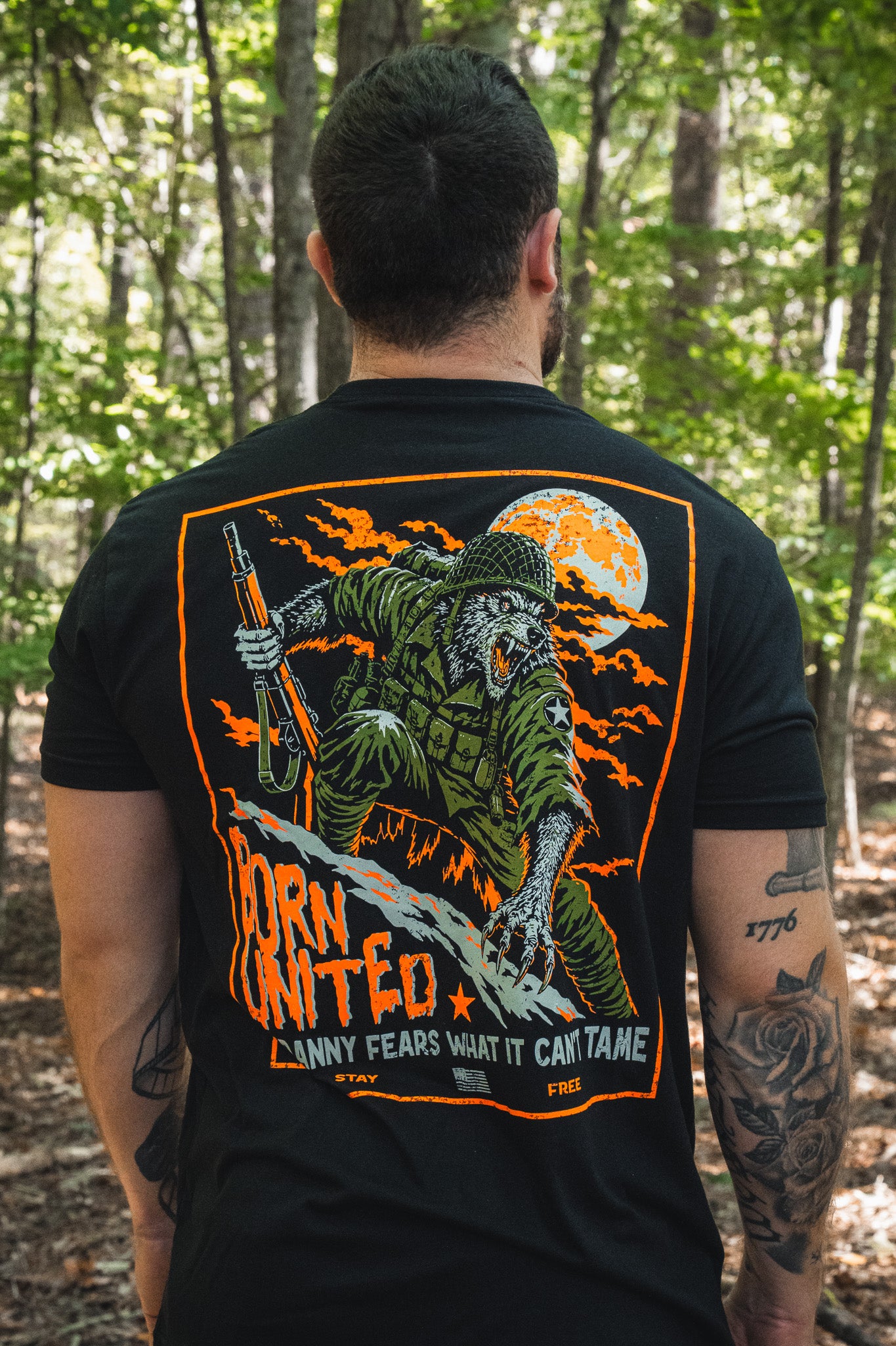 Back view of a person wearing a black Full Moon Tee featuring a tactical werewolf soldier holding a rifle in front of a full moon, with the text BORN UNITED and ANY FEARS WHAT IT CANT TAME.