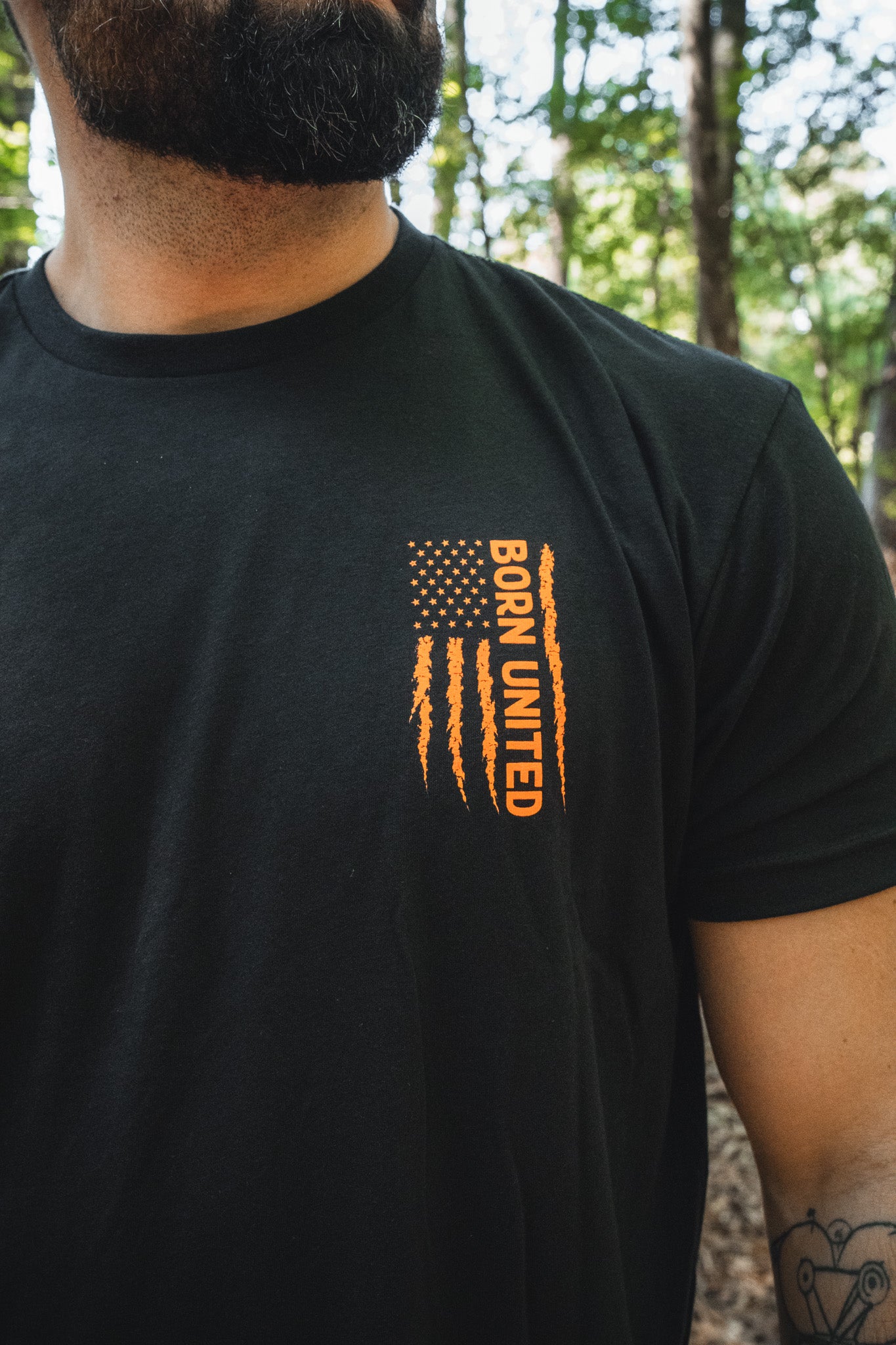 A man wears a black tactical Full Moon Tee featuring an orange, distressed American flag graphic with the text BORN UNITED printed vertically.