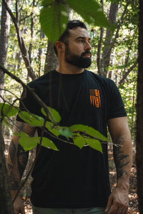 A bearded man stands in a forest wearing a black Full Moon Tee featuring a tactical werewolf and a distressed American flag graphic.