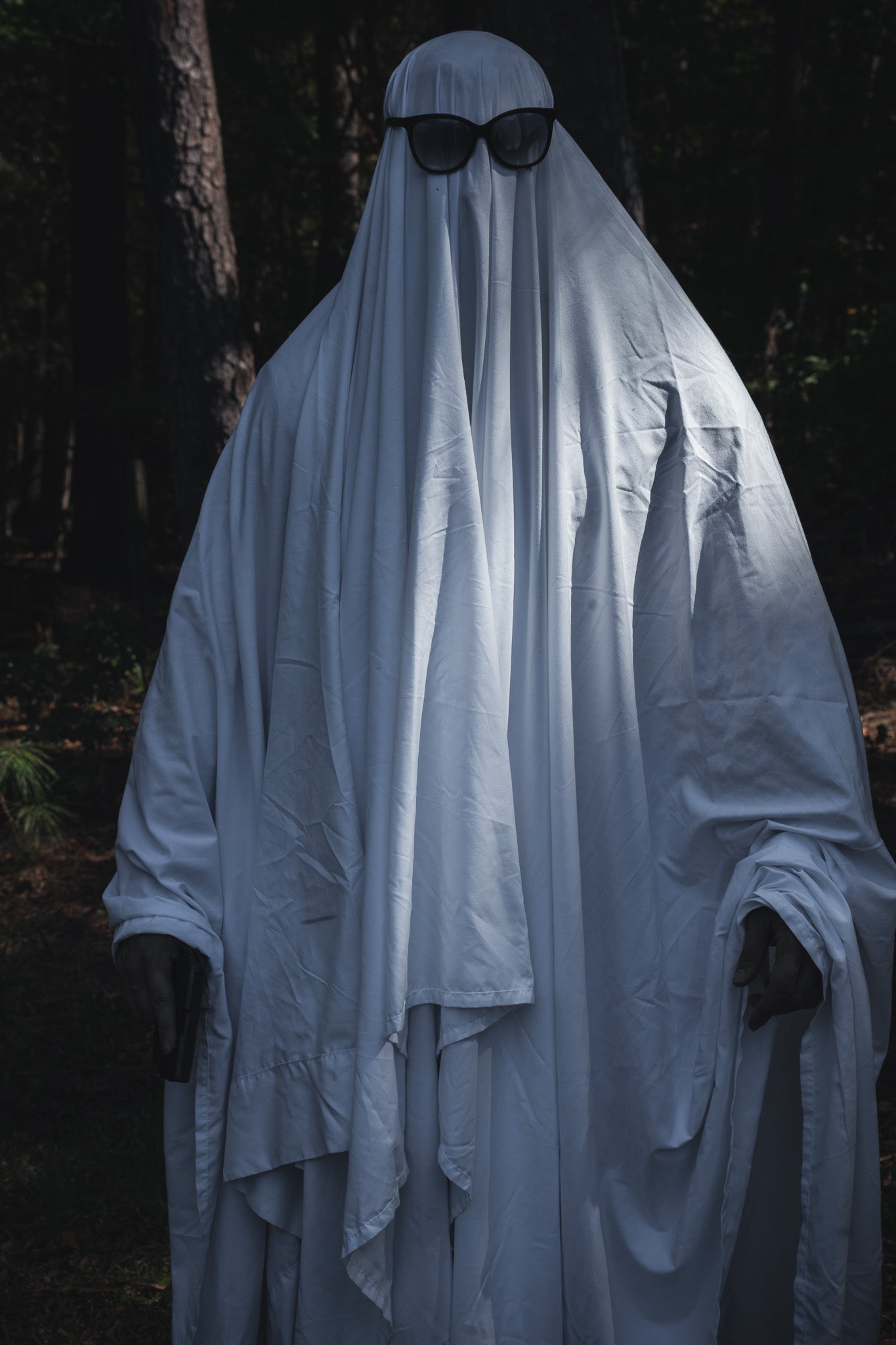 A person wearing a Ghost Ops Tee under a Dead But Deployable ghost costume made from a white sheet and black sunglasses, standing in a wooded area.
