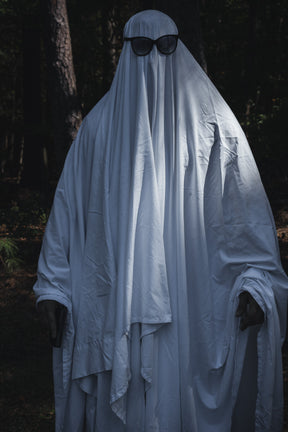 A person wearing a Ghost Ops Tee under a Dead But Deployable ghost costume made from a white sheet and black sunglasses, standing in a wooded area.