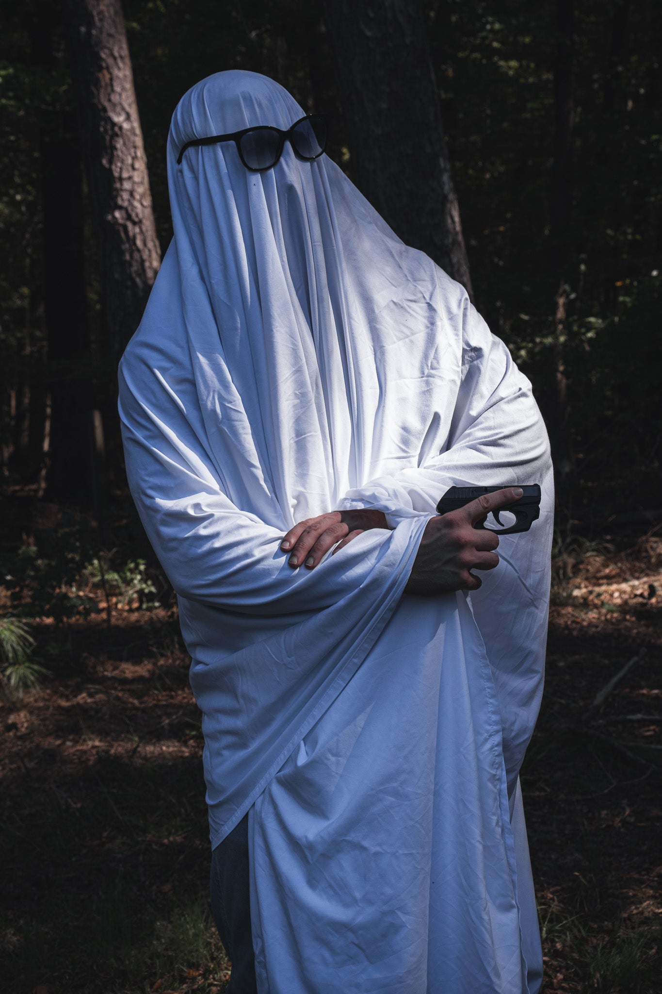A person wearing a Ghost Ops Tee with a Dead But Deployable ghost costume, sunglasses, and a handgun in the woods.