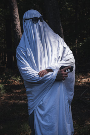 A person wearing a Ghost Ops Tee with a Dead But Deployable ghost costume, sunglasses, and a handgun in the woods.