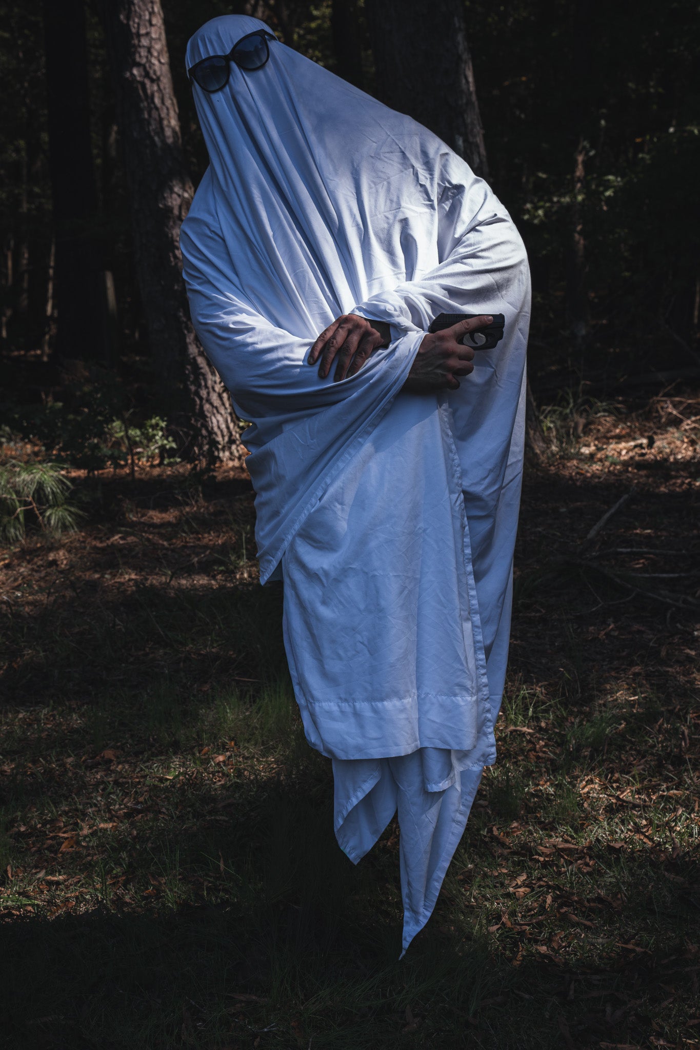 A person wearing a Ghost Ops Tee in a Dead But Deployable ghost costume with a white sheet and sunglasses stands in a forest while holding a handgun.