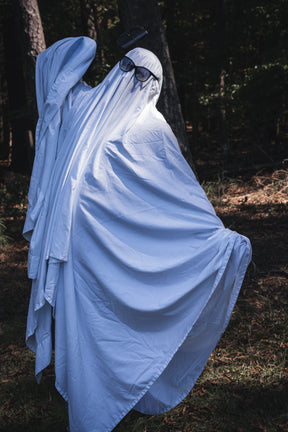 A person wearing a Ghost Ops Tee in a Dead But Deployable ghost costume with black-rimmed glasses and a handgun tucked into the sheet, standing in a wooded area.