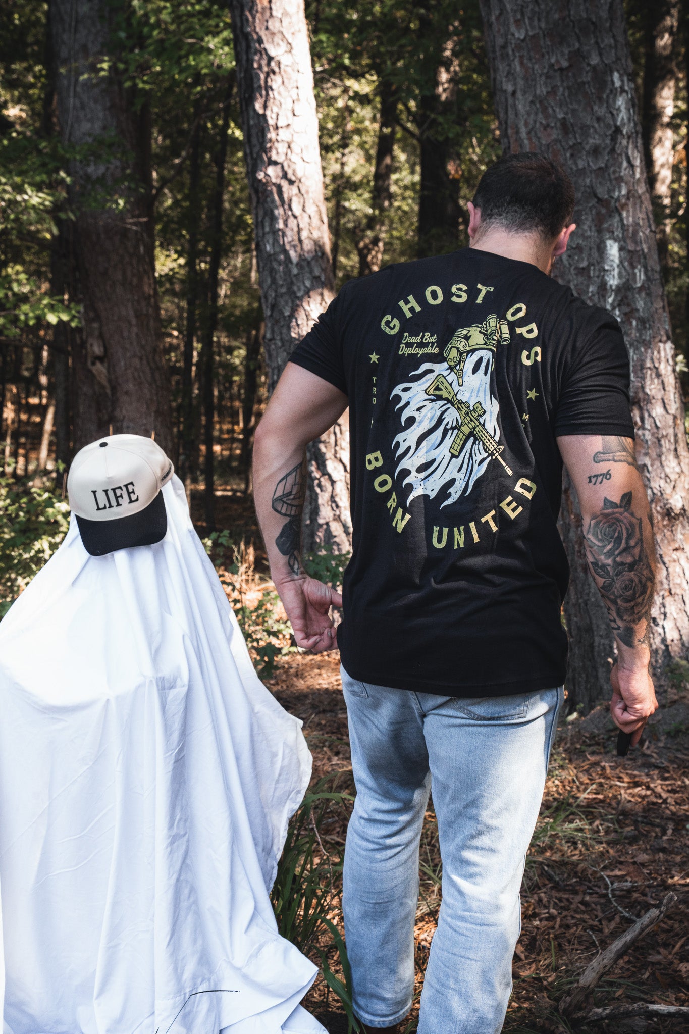 A man wearing a Ghost Ops Tee walks in the woods next to a person dressed as a ghost wearing a baseball cap that says LIFE.