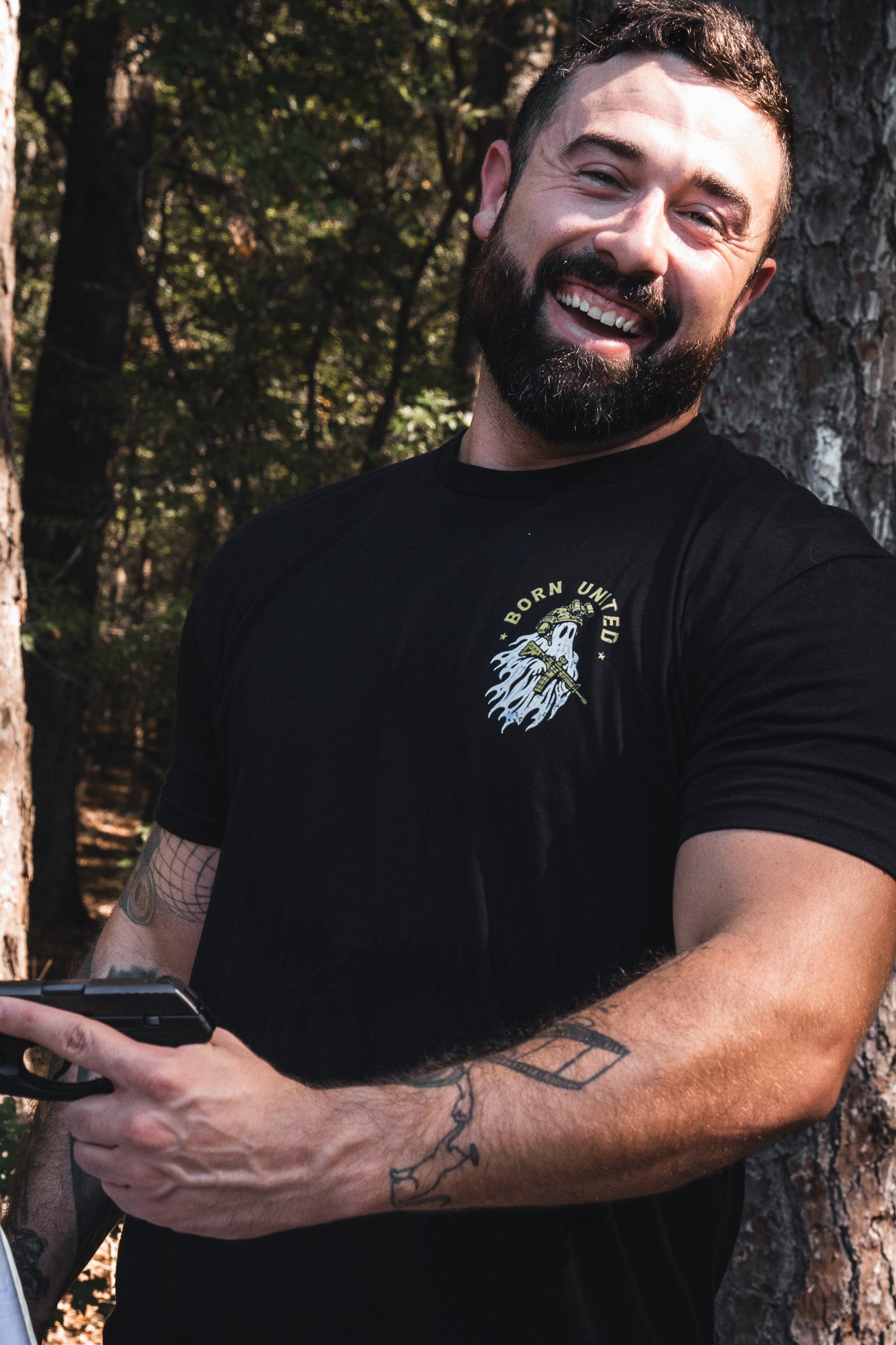 A bearded man wearing a black Born United Private Label Ghost Ops Tee holds a handgun.