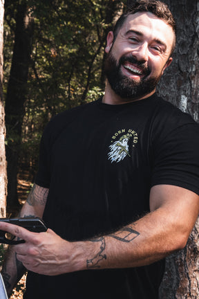 A bearded man wearing a black Born United Private Label Ghost Ops Tee holds a handgun.