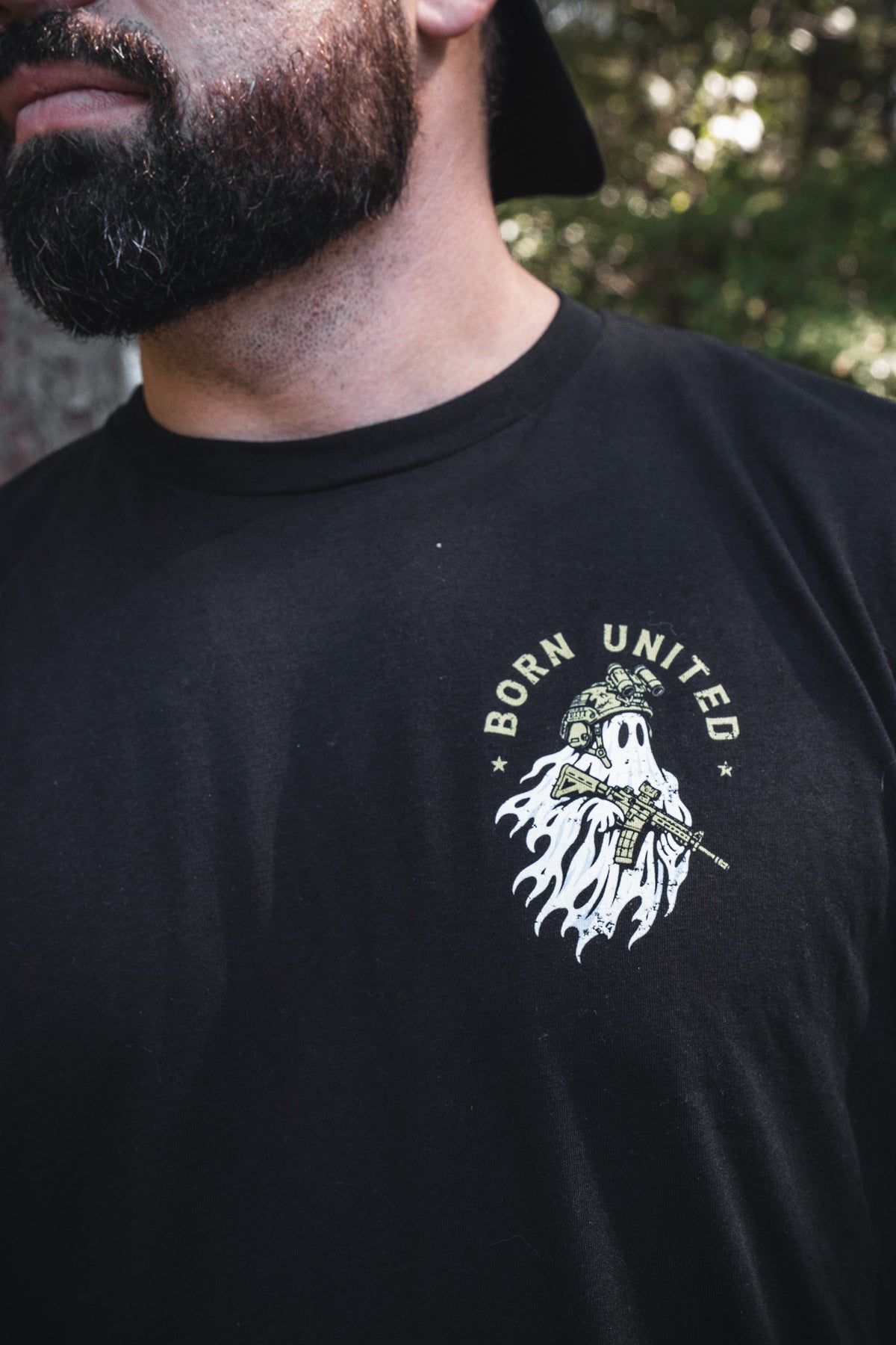 A man wearing a black Ghost Ops Tee featuring a graphic of a ghost in a tactical helmet holding a rifle, with the words BORN UNITED arched above it.