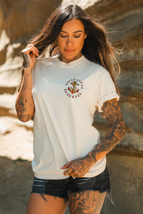 A woman with long brown hair and tattooed arms stands against rocky terrain, wearing the Relentless Betrayal Into the Storm Premium Ivory T-Shirt and black denim shorts. She looks down, gently touching her hair.