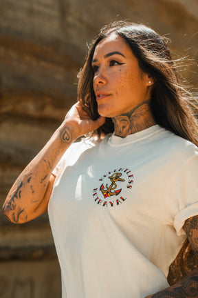A woman with long hair and tattoos stands outdoors wearing the Relentless Betrayal Into the Storm Premium Ivory T-Shirt, made from jersey cotton and featuring an anchor graphic with the words No Titles, No Betrayal.