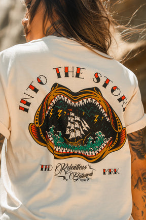 Someone wears the Relentless Betrayal Into the Storm Premium Ivory T-Shirt, featuring a vivid back graphic of an open-mouthed sea creature with a ship inside, waves, lightning, and “INTO THE STORM” text above.