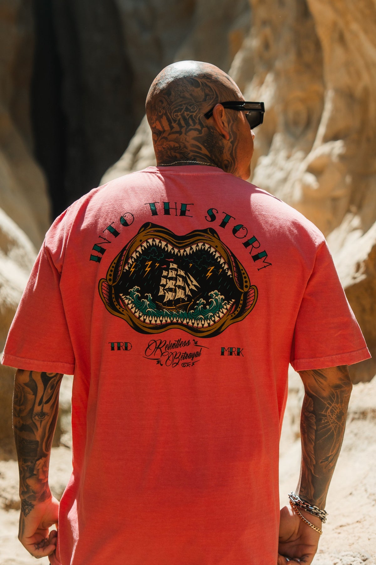 A tattooed man in sunglasses wears the Relentless Betrayal Into the Storm Premium Watermelon T-Shirt, featuring a sailing ship and “INTO THE STORM” on the back, as he stands outdoors in a rocky, sunlit area.