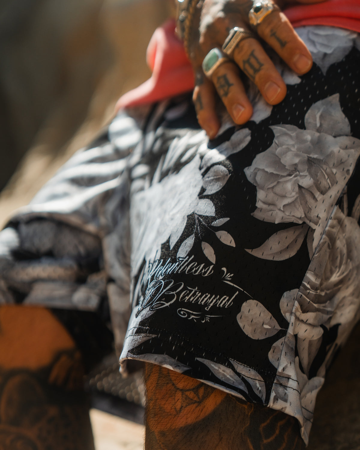 A tattooed hand with rings rests on Relentless Betrayal’s Premium Mesh Shorts | 5–Beauty in Darkness, featuring black and white floral patterns and cursive text. The unisex shorts are styled with a red shirt for a comfortable, bold look.