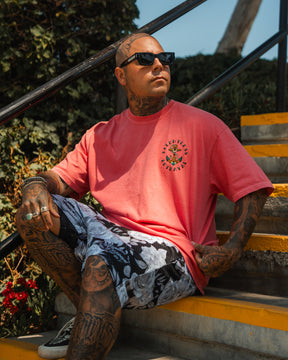 A tattooed man in sunglasses, a pink T-shirt, and Relentless Betrayals Premium Mesh Shorts | 5- Beauty in Darkness sits on outdoor stairs amidst greenery, sunlight creating dramatic shadows around him.