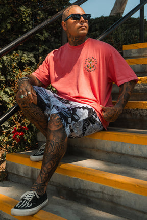 A tattooed man in sunglasses and patterned shorts sits on outdoor steps, wearing the Relentless Betrayal Into the Storm Premium Watermelon T-Shirt, made of jersey cotton, as he looks off to the side on a sunny day.