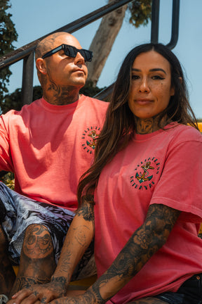 A tattooed man and woman sit on outdoor steps in matching Relentless Betrayal Into the Storm premium watermelon t-shirts with an anchor graphic. The man wears sunglasses and looks up; the woman smiles slightly at the camera.