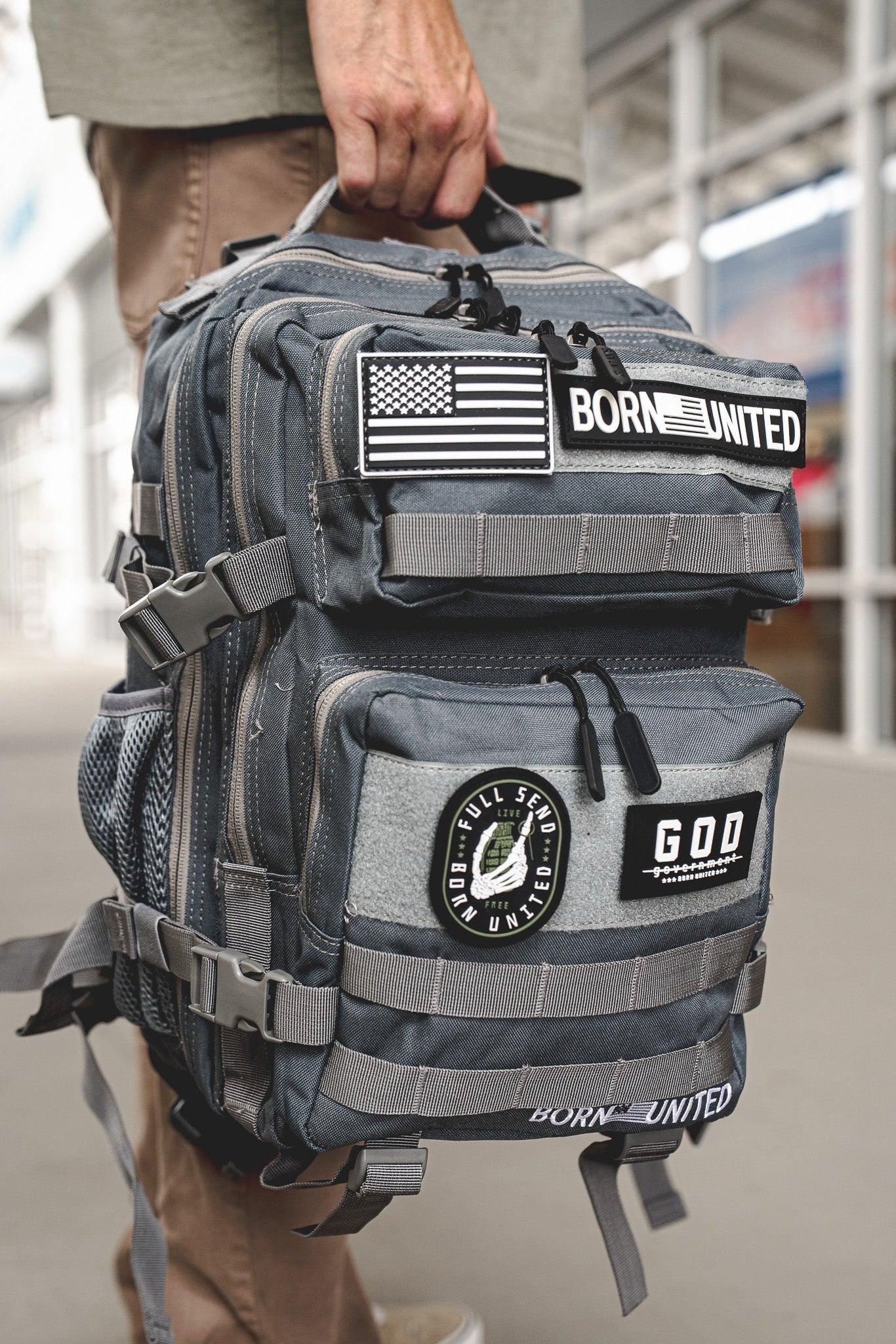 A person holds the Born United Private Label Standard Tac-Pac Ultimate Bundle backpack with patches—U.S. flag, BORN UNITED, and a GOD FULL SEND UNITED circle—while wearing a Born United tee outdoors in an urban area.